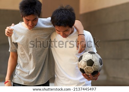 Good game. Two asian boys walking with their arms around eachother holding a soccer ball.