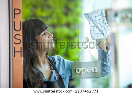 Portrait of smiling owner cleaning the door at her cafe with open signboard. Coffee owner in coffee shop to welcome customer and open the coffee shop in morning.