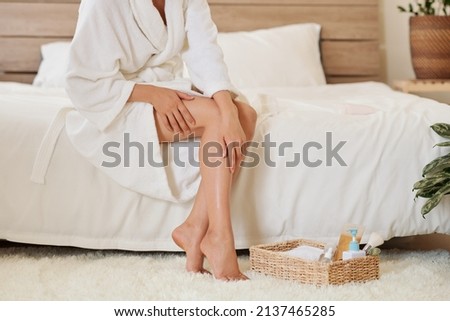 Young woman in bathrobe sitting on bed and applying body oil after shower Royalty-Free Stock Photo #2137465285