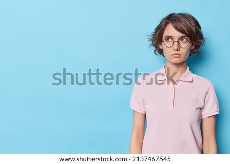 Waist up shot of young brunette woman concentrated away with serious expression feels confident thinks about something wears round spectacles casual t shirt isolated over blue background copy space
