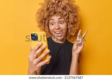 Indoor shot of happy positive curly haired woman sticks out tongue makes peace gesture takes selfie via mobile phone makes photo of herself wears casual black t shirt isolated over yellow background.