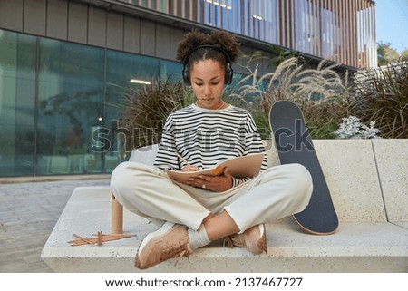 Horizontal shot of skilled creative curly woman makes drawings in notepad sits crossed legs listens pleasant music via headphones poses outdoors in urban setting rests after riding skateboard