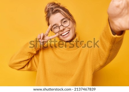 Glad young woman makes peace gesture over eye smiles broadly takes selfie wears round spectacles and casual jumper isolated over yellow background stays always positive. Body language concept