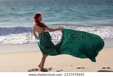 Full length portrait of  red haired woman wearing a  beautiful  long green  silk toga gown. Standing  pose with gestural hands at  ocean beach landscape background.
