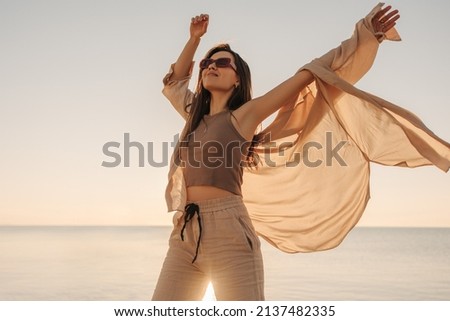 Cheerful young fair-skinned lady waving her arms above head against background of sea. Brown-haired woman with straight hair wearing sunglasses. Cozy beach atmosphere, summer concept.