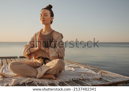 Relaxed young caucasian woman sitting on seashore practices yoga without stress. Model with dark topknot on her head with eyes closed. Cozy beach atmosphere, summer concept. Royalty-Free Stock Photo #2137482341