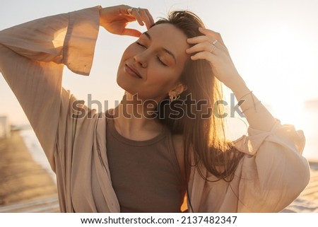 Close-up of young caucasian relaxed woman touching her hair in bright sunlight. Brown-haired girl with closed eyes, wearing brown T-shirt and blouse on top. Good mood, fashion trends Royalty-Free Stock Photo #2137482347