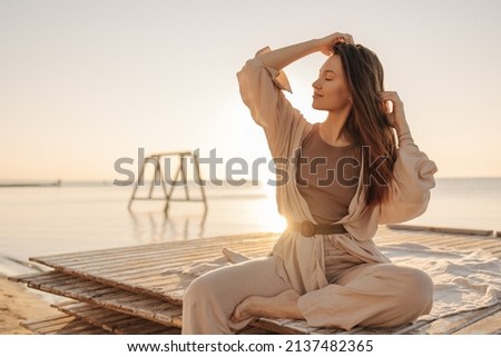 Smiling young caucasian girl, in light suit, trains on beach by sea. Woman with straight dark hair closed her eyes and turned head to side. Rest time concept. Royalty-Free Stock Photo #2137482365