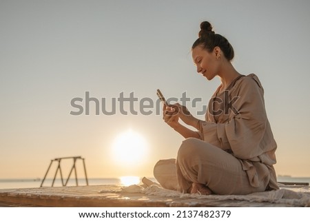 Profile photo of pretty cheerful young woman sitting cross-legged with phone against the sky. Caucasian lady with bun of hair communicates in hundreds of networks with friends, dressed casual clothes.