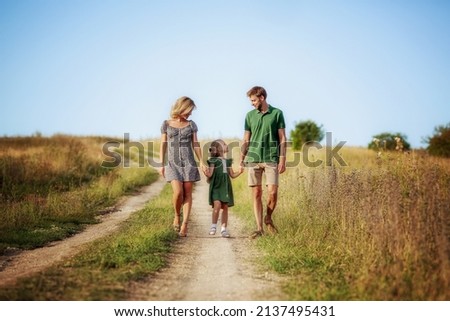 A happy family walks hand in hand along a field road. Parents hold their daughter by the hand. Spend time together. Walk outdoors  Royalty-Free Stock Photo #2137495431