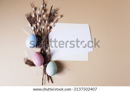 Multicolored eggs on a beige background next to willow branches and a white sheet for text. Easter picture with copy space.