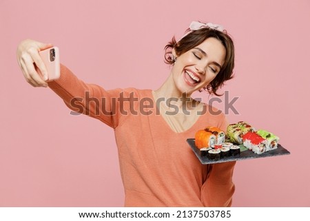 Young fun happy woman wear sweater hold in hand makizushi sushi roll served on black plate traditional japanese food doing selfie shot on mobile cell phone isolated on plain pastel pink background.