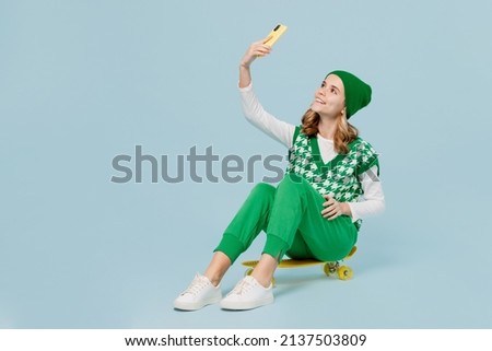 Full size cheery young brunette girl teen student wears checkered green vest hat sit on skateboard doing selfie shot on mobile cell phone isolated on plain pastel light blue background studio portrait
