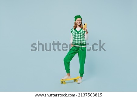 Full size body length young brunette girl teen student wears checkered green vest hold in hand use mobile cell phone stand on skateboard isolated on plain pastel light blue background studio portrait