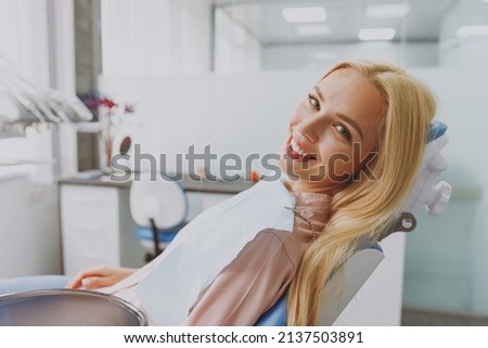 Close up young smiling happy blonde woman 20s covered by napkin sitting at dental office chair indoor light modern cabinet waiting stomatologist for oral procedure. Healthcare caries enamel treatment. Royalty-Free Stock Photo #2137503891
