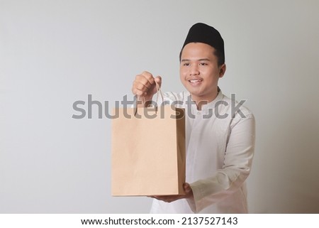 Portrait of Asian muslim man in white koko shirt with skullcap giving craft paper shopping bag with gift inside. Isolated image on gray background
