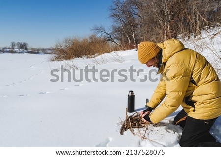 A tourist in a yellow down jacket makes a fire in the snow. Survival in the forest, bushcraft, a warm hat on your head, a winter hike to equip camp, cook food. High quality photo