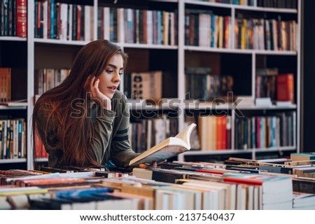 Beautiful young woman buying books at a bookstore and reading one. Geeky woman reading a book with a bookshelves in background. Royalty-Free Stock Photo #2137540437