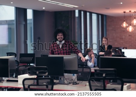 A young man with an Afro haircut stands in a modern office surrounded by computers