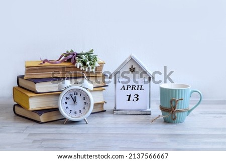 Calendar for April 13: a decorative house with the name of the month April in English, number 13, a stack of books, a bouquet of snowdrops on them, an alarm clock, a cup, a gray background