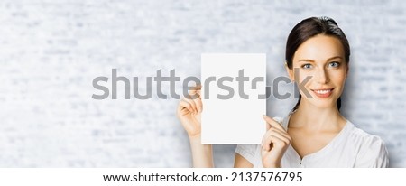 Portrait image of cheerful smiling woman in white cloth, holding showing demonstrate mock up paper signboard. Business and advertising concept. Copy space empty area. White bricks wall.