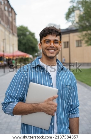 Authentic portrait of young handsome Indian man wearing stylish eyeglasses standing on the street. Smiling asian student holding laptop looking at camera in university campus. Education concept  Royalty-Free Stock Photo #2137584357