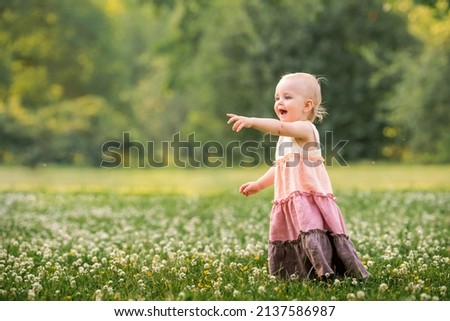little blue-eyed girl walks in a meadow with white clover in summer in a long vintage linen dress, points with her finger.
