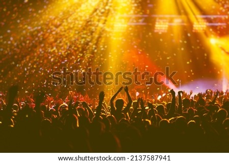 A crowded concert hall with scene stage orange and yellow lights, rock show performance, with people silhouette, colourful confetti explosion fired on dance floor air during a concert festival
