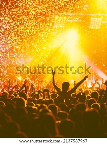 A crowded concert hall with scene stage orange and yellow lights, rock show performance, with people silhouette, colourful confetti explosion fired on dance floor air during a concert festival
 Royalty-Free Stock Photo #2137587967