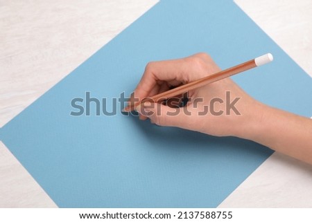 Woman drawing on light blue paper with pastel pencil at white wooden table, top view
