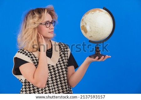 Learning about the globe. Geography concept. Young teenage girl in glasses holding artificial globe with one hand over blue background. Studio shot. High quality photo