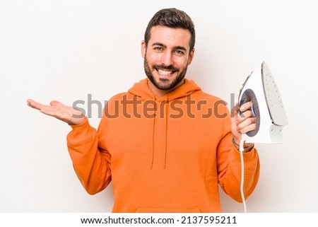 Young caucasian man holding an iron isolated on white background showing a copy space on a palm and holding another hand on waist.