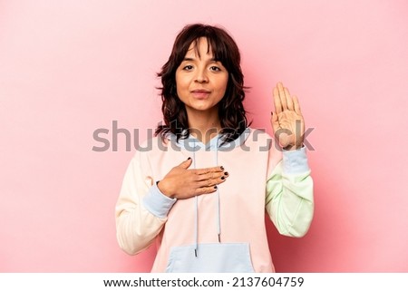 Young hispanic woman isolated on pink background taking an oath, putting hand on chest.