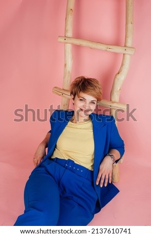 Elegant woman in a blue classic suit and yellow blouse on a pink background. Yellow and blue clothes. Yellow shoes and blue trouser suit. Pink background and wooden ladder