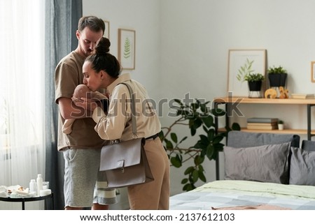 Young affectionate female kissing cute baby son on hand of her husband in the morning while saying goodbye and leaving for work Royalty-Free Stock Photo #2137612103
