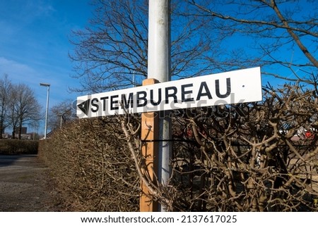 A white sign against a deep blue sky with black lettering that reads "Stembureau" (polling station) directs voters to the correct location.