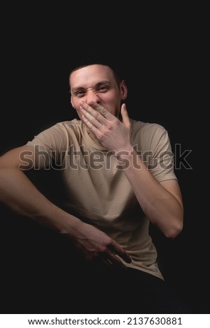 Handsome young man posing over black background. Studio photo with one light source. Smiling guy cheerfully posing in the studio. The guy covers his face with his hand, yawning.