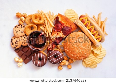 Junk food table scene. Collection scattered over a white marble background. Mixture of take out and fast foods. Pizza, hamburgers, french fries, chips, hot dogs, sweets. Overhead view. Royalty-Free Stock Photo #2137640529