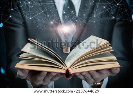A man in a suit shows an open textbook and a burning light bulb on a dark background. Royalty-Free Stock Photo #2137644829