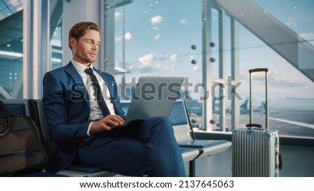 Busy Crowded Airport Terminal: Businessman Uses Laptop, Waiting for a Flight. Traveling Entrepreneur Working Online On Computer Sitting in a Boarding Lounge of Airline Hub with Airplanes