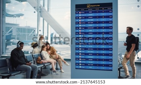 Airport Terminal: Young Man Looking at Arrival and Departure Information Display Looking for His Flight. Backgrond: Diverse Crowd of People Wait for their Flights in Boarding Lounge of Airline Hub Royalty-Free Stock Photo #2137645153