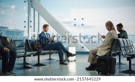 Busy Airport Terminal: Handsome Businessman Uses Smartphone While Waiting for His Flight. People Sitting in a Boarding Lounge of Big Airline Hub with Airplanes Departing and Arriving Royalty-Free Stock Photo #2137645229