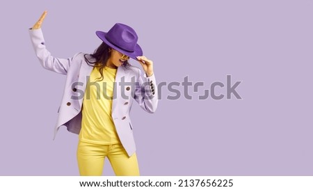 Cheerful charismatic stylish woman having fun and dancing isolated on pastel purple background. Cool smiling woman in fashionable bright clothes dances holding hat near copy space. Banner.