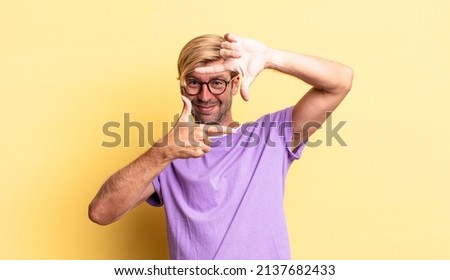 handsome blond adult man feeling happy, friendly and positive, smiling and making a portrait or photo frame with hands