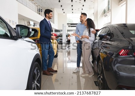 Wealthy middle eastern young couple having conversation with good-looking sales assistant at luxury auto showroom, looking for new car, gesturing and smiling, full length photo, panorama Royalty-Free Stock Photo #2137687827