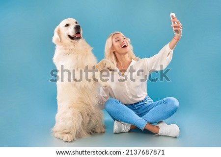 Excited beautiful caucasian blonde woman taking selfie with her happy dog, posing holding gadget looking at device camera, playful pet standing on rear hind legs isolated on blue studio background