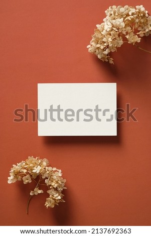 Blank white paper card mockup with dried flowers on marsala color background. Flat lay, top view, copy space.
