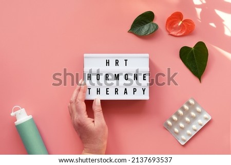 Text HRT Replacement Therapy on light box in hand. Menopause, hormone therapy concept. Estrogen replacement therapy awareness design. Pink background with exotic leaves, flowers, pills, estrogene gel. Royalty-Free Stock Photo #2137693537