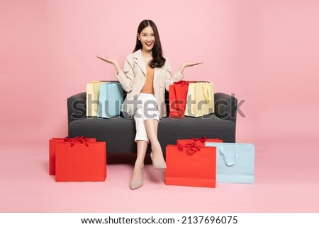 Full length portrait of Excited Asian woman sitting on sofa with shopping bags isolated on pink background, Shopper or shopaholic concept Royalty-Free Stock Photo #2137696075