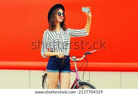Beautiful happy smiling young woman taking selfie by smartphone with bicycle wearing a shirt, shorts, black round hat on red background